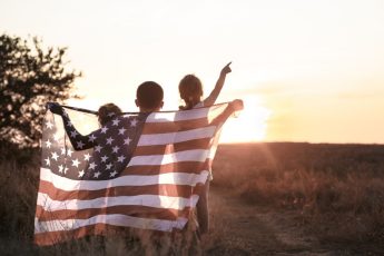 Niños pequeños con una bandera estadounidense envuelta a sus espaldas al atardecer.