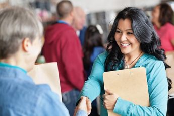 Young woman holding a clipboard while shaking hands with another woman.