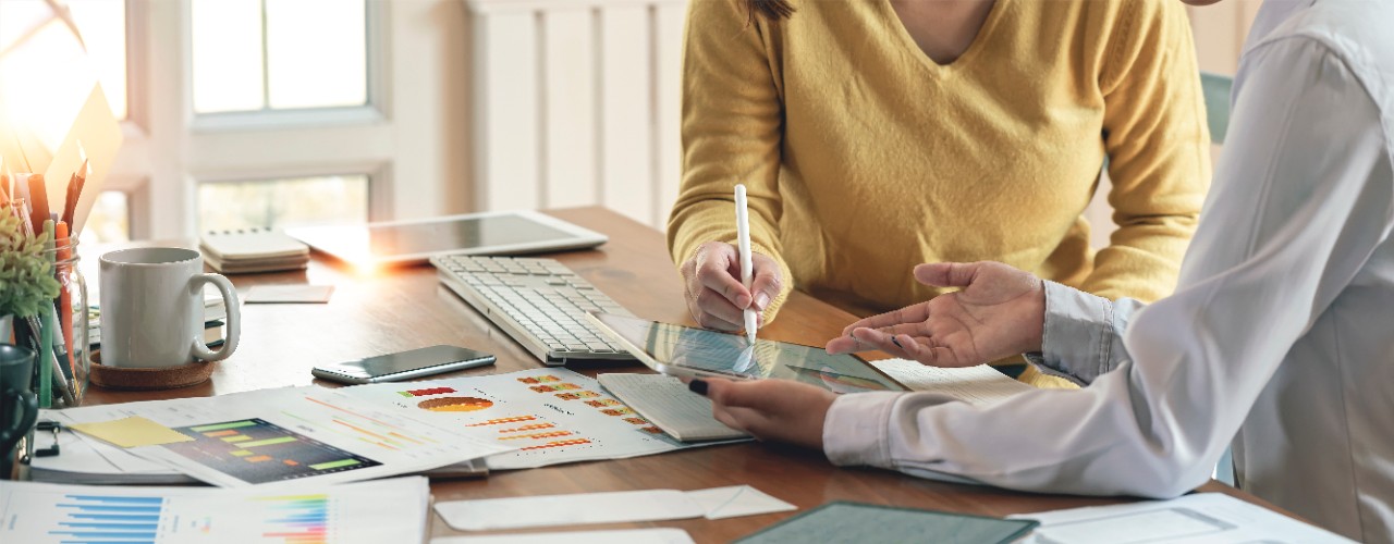 The image shows two people sitting at a desk working together with a digital tablet. Datasheets are laid out in front of them on the desk.