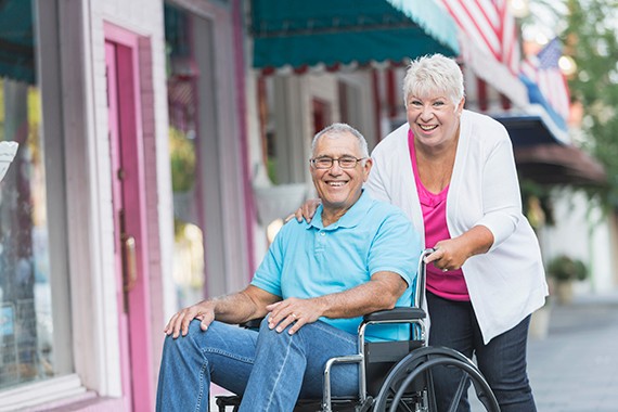 A senior couple on the sidewalk in front of stores. The man is sitting in a wheelchair and his wife is behind him with her hand on his shoulders. They are smiling and looking at the camera.