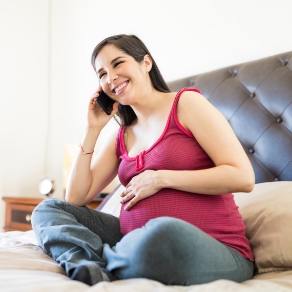 Cheerful woman, sitting on a bed, chatting on call while touching pregnant tummy.