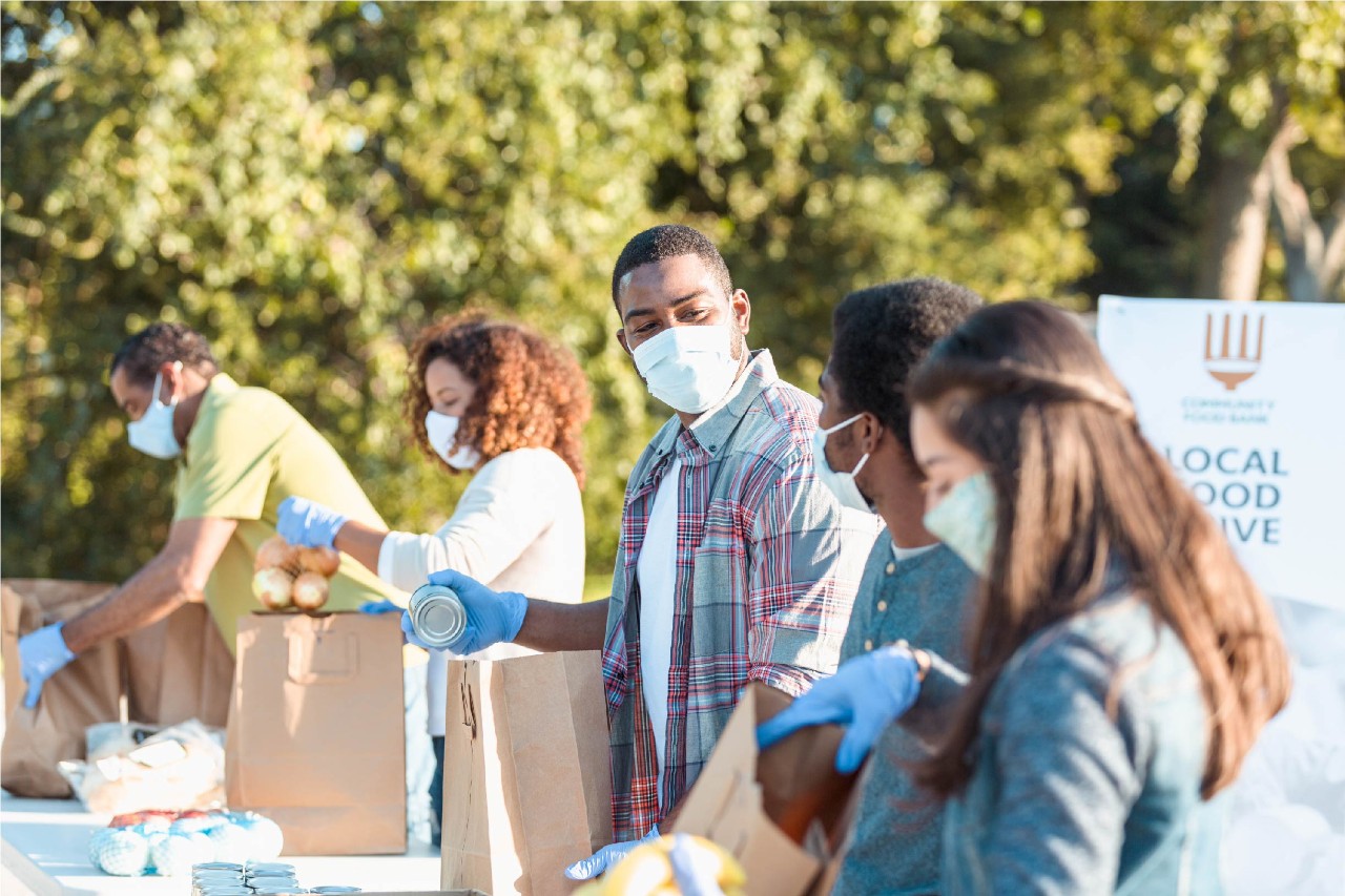 An image of a male volunteer places a canned food item into a paper bag at an outdoor food bank. He and other volunteers are wearing protective face masks as they are volunteering during the coronavirus pandemic.
