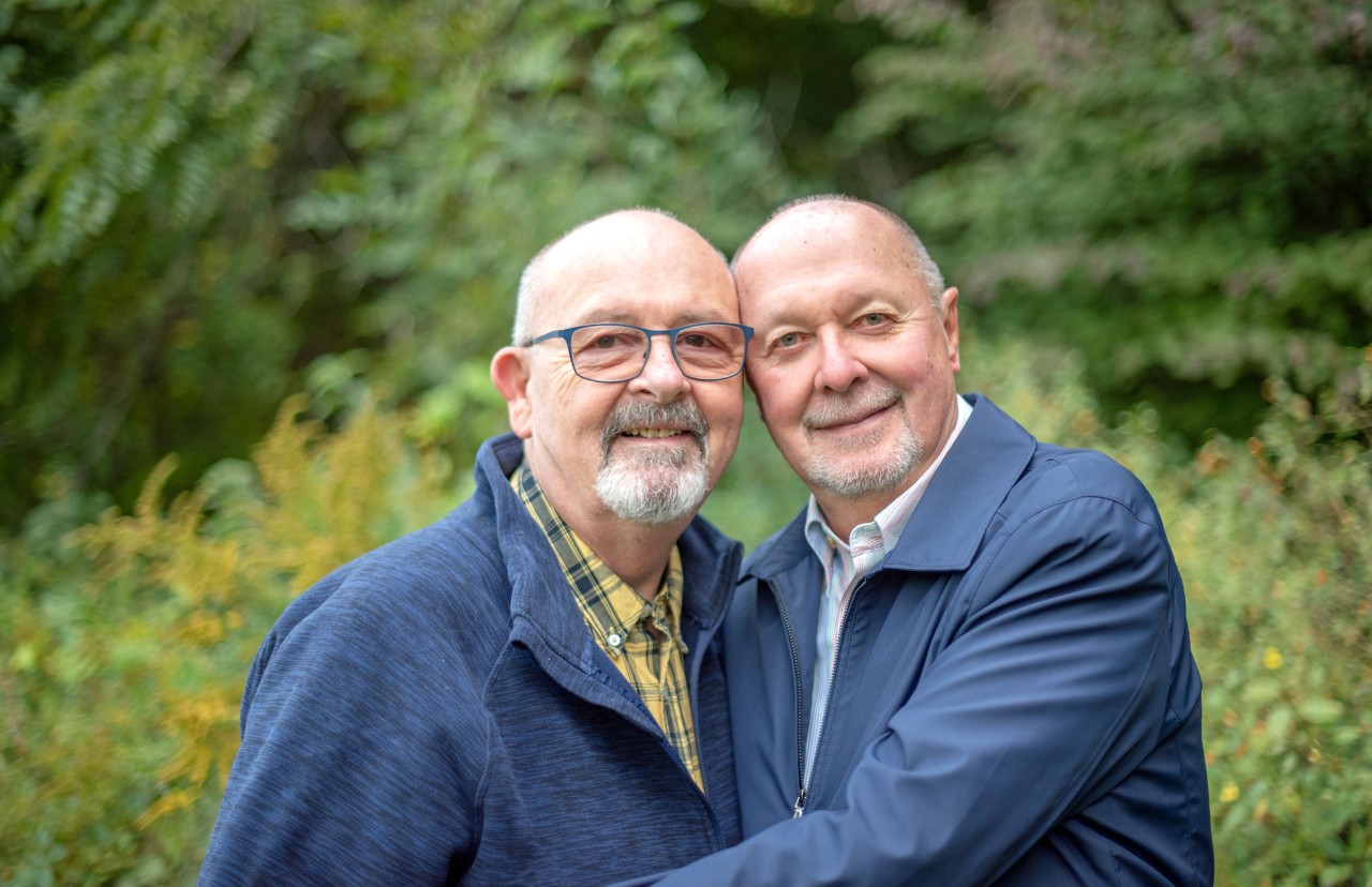 Married, elderly gay male couple embrace each other.