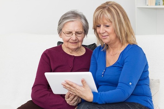 Mama de edad avanzada con su hija adulta sentadas en el sofa revisando informacion en una tableta electronica.