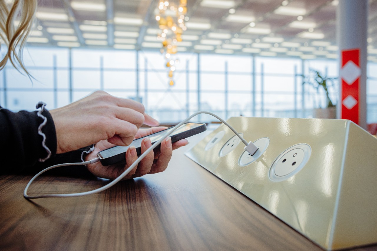 Primer plano de una mujer con cabello rubio, vestida con un jersey negro, usando su teléfono inteligente mientras lo carga a través de un cable USB blanco en una estación de carga pública instalada en una mesa de madera dentro de una terminal de aeropuerto, ventana de la terminal en el fondo, enfoque en primer plano, horizontal